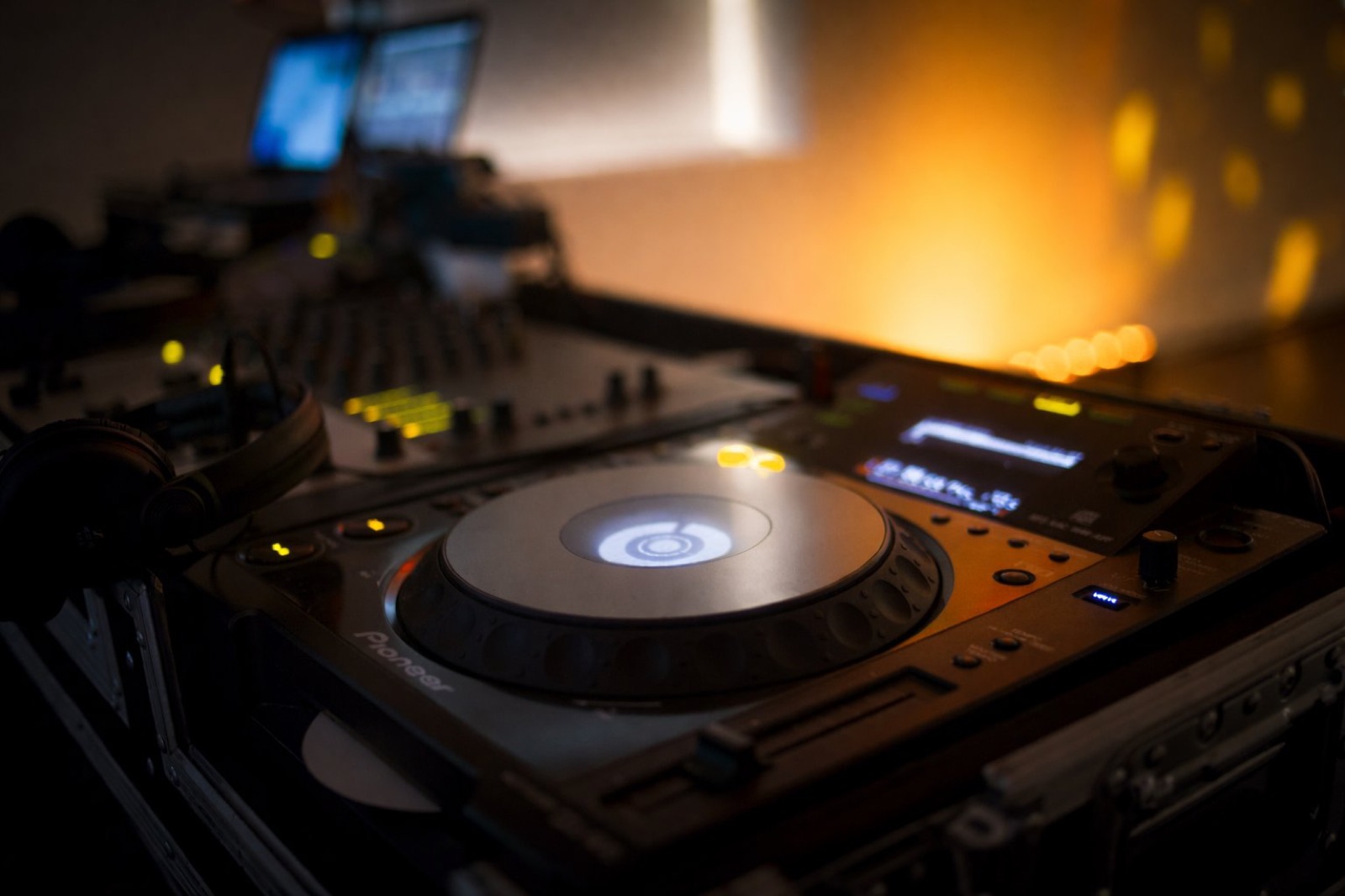 Close-up of a Pioneer CDJ deck glowing warmly, with a mixing console and laptops softly out of focus behind it