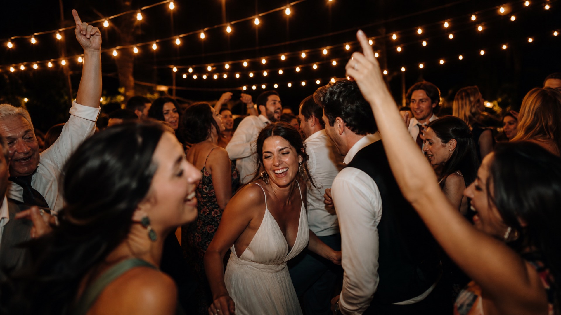 Guests laughing and dancing together under warm string lights on a night-time dancefloor