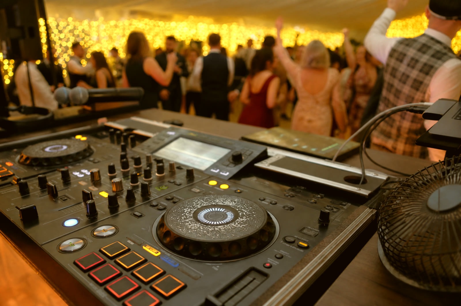 DJ controller in the foreground with wedding guests dancing under warm string lights in the background